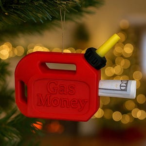 May include: A red gas can Christmas ornament with the words "Gas Money" printed on the front. A rolled-up US $100 bill is inserted into the opening. The gas can has a yellow spout and is hanging from a Christmas tree.