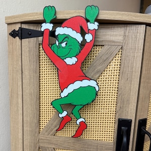 May include: A cartoon Grinch figure in a red Santa hat and coat with white trim, green skin, and red shoes, clinging to a wooden cabinet door. The cabinet has a woven panel and black hardware.