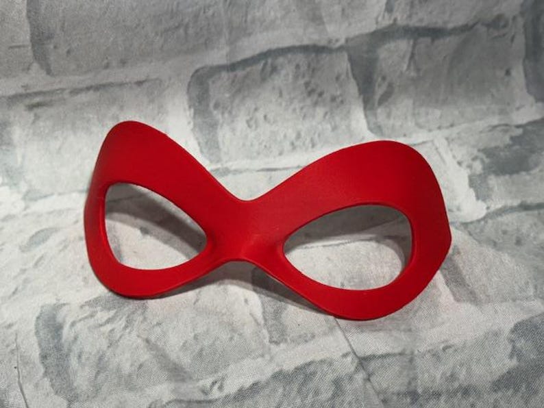 Puede incluir: Una m&aacute;scara de ojos roja vibrante con una forma distintiva, con dos grandes aberturas para los ojos conectadas por un puente curvo. La m&aacute;scara est&aacute; hecha de un material liso y se coloca sobre un fondo texturizado de color claro.