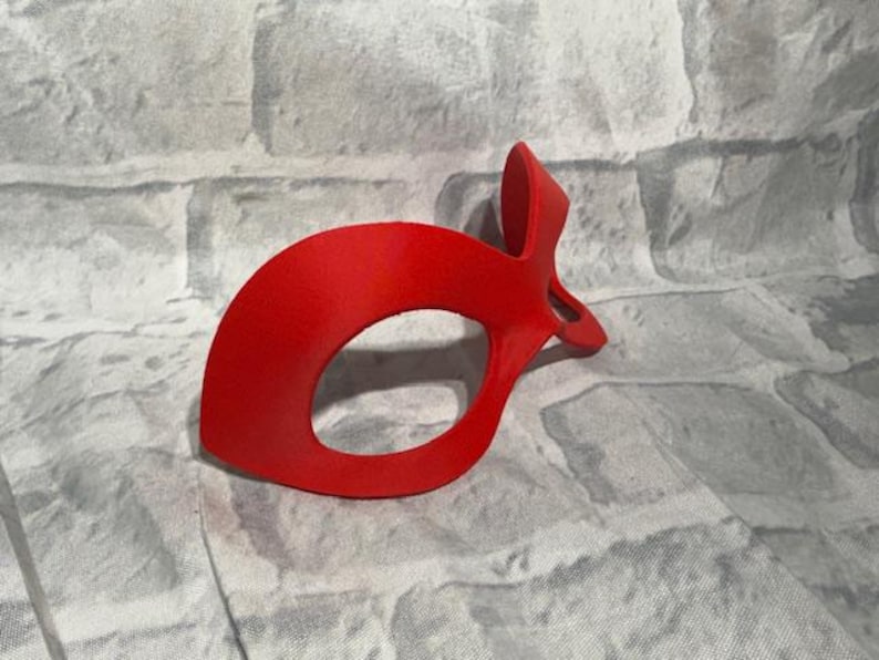 Puede incluir: Una m&aacute;scara de ojos roja brillante con un dise&ntilde;o curvo y una parte superior puntiaguda. La m&aacute;scara tiene una gran abertura ovalada para el ojo y est&aacute; hecha de un material liso y s&oacute;lido. Probablemente para disfraces o uso teatral.