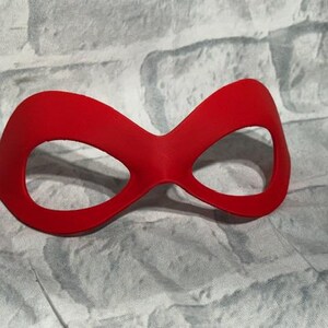 Puede incluir: Una m&aacute;scara de ojos roja brillante con una forma curva y dos grandes aberturas para los ojos. La m&aacute;scara est&aacute; hecha de un material liso y probablemente se utiliza para disfraces o teatro. El fondo es una superficie texturizada de color gris claro.