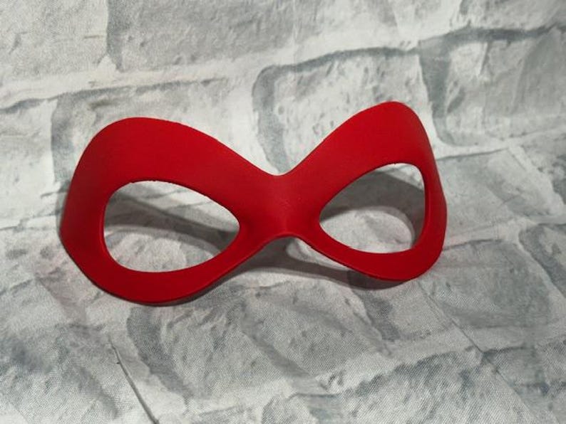Puede incluir: Una m&aacute;scara de ojos roja brillante con una forma curva y dos grandes aberturas para los ojos. La m&aacute;scara est&aacute; hecha de un material liso y probablemente se utiliza para disfraces o teatro. El fondo es una superficie texturizada de color gris claro.