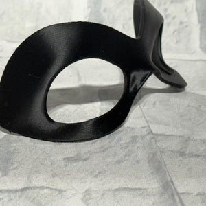 Puede incluir: Una m&aacute;scara de ojos negra con un dise&ntilde;o curvo y una abertura ovalada para los ojos. La m&aacute;scara tiene una textura suave, similar al sat&eacute;n, y una peque&ntilde;a extensi&oacute;n puntiaguda en la parte superior. La m&aacute;scara es probablemente para disfraces o para dormir.