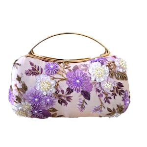 May include: A rectangular clutch with a gold handle, adorned with purple and white floral embellishments. The flowers are intricately detailed with beads and sequins, set against a pale pink background.