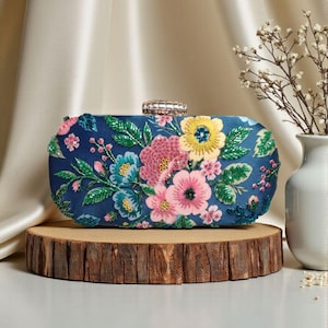 Può includere: Una pochette blu navy con un motivo floreale di fiori rosa, gialli e blu. La borsa rettangolare ha una chiusura argentata ed è impreziosita da perline. La pochette è esposta su una fetta di legno.