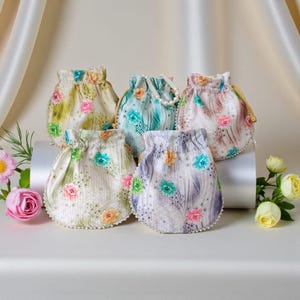 Op de afbeelding: Vijf bloemen trekkoordtassen in verschillende pastelkleuren, waaronder geel, blauw en roze. Elke tas heeft een delicaat bloemenpatroon en is versierd met kleine parels langs de onderrand. Sommige tassen hebben parelhandvatten.