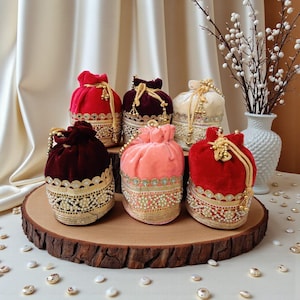Può includere: Sei pochette in velluto con coulisse nei toni del rosso, bordeaux e pesca, con finiture dorate e perlate. Le pochette sono esposte su una fetta di legno, con un vaso di fiori bianchi sullo sfondo.