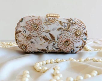 Könnte beinhalten: Eine ovale Clutch mit floralem Design. Die Clutch ist weiß mit rosa und goldfarbener Stickerei, mit Perlenblumen und goldenen Blättern. Sie hat einen goldenen Verschluss und liegt vor einem weißen Hintergrund mit einer Perlenkette.