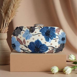 Peut inclure: Pochette blanche ornée de broderies florales bleues et de perles. Le sac rectangulaire est doté d'un fermoir orné de bijoux et décoré de fleurs bleu foncé et bleu clair, avec des feuilles marron et noires.