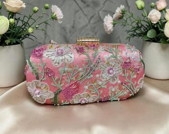 Könnte beinhalten: Rechteckige, rosa Clutch mit floralen Stickereien in Weiß und Rosa, grünen Stielen und Perlenakzenten. Die Tasche hat einen goldenen Verschluss und steht vor einem neutralen Hintergrund, flankiert von Blumenarrangements.