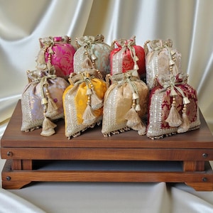 Puede incluir: Un surtido de bolsas decorativas con cordón en varios colores, incluyendo rosa, dorado, rojo y amarillo. Cada bolsa presenta adornos dorados ornamentados, borlas y un cierre de cordón. Las bolsas se exhiben sobre una plataforma de madera oscura.