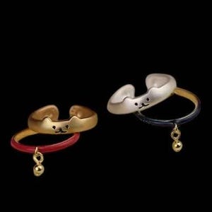 May include: Two adjustable rings with a cat face design. One ring is gold with a red band and a gold charm. The other ring is silver with a black band and a gold charm.