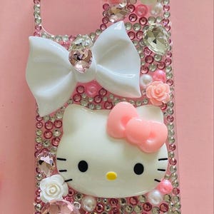 May include: Pink phone case decorated with rhinestones, white bow, and Hello Kitty character. The case features pink and white roses, pearls, and large crystal accents. The case is designed for a smartphone.