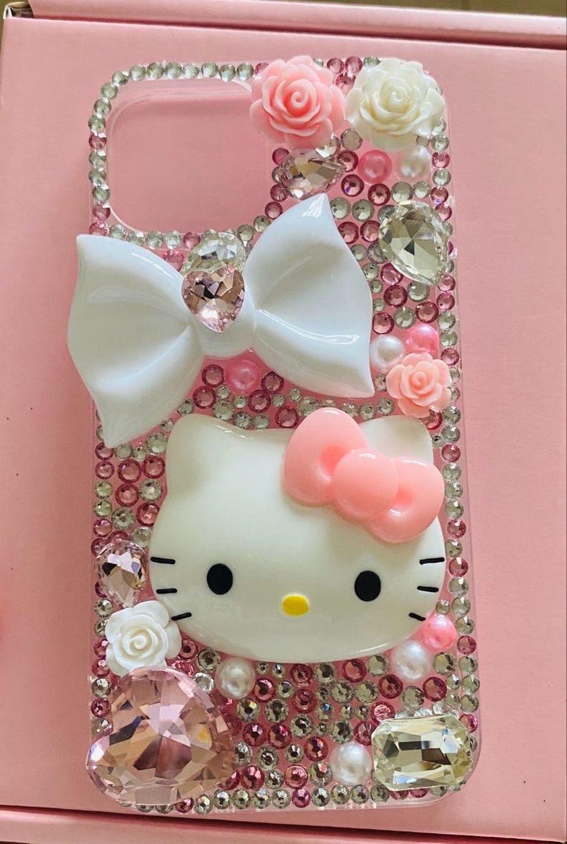 May include: Pink phone case decorated with rhinestones, white bow, and Hello Kitty character. The case features pink and white roses, pearls, and large crystal accents. The case is designed for a smartphone.