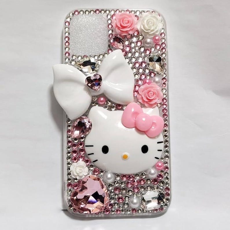 May include: A decorative phone case featuring a white Hello Kitty design with a pink bow, surrounded by pink and clear rhinestones, white pearls, and pink roses. The case has a silver glitter background and a large white bow.