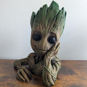 May include: A detailed, green and brown sculpture of a character with a tree-like appearance, featuring large black eyes and a gentle smile. The figure rests its head on its hand, with a thoughtful expression. The sculpture is likely a decorative item.