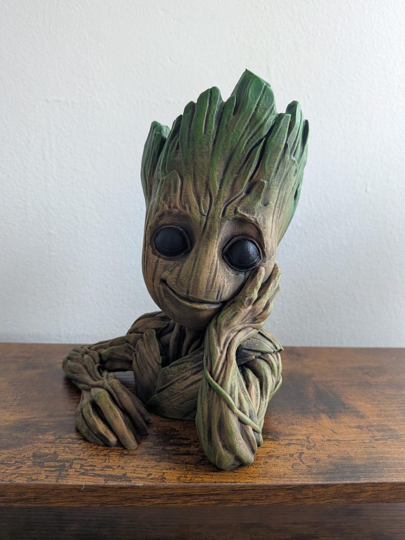 May include: A detailed, green and brown sculpture of a character with a tree-like appearance, featuring large black eyes and a gentle smile. The figure rests its head on its hand, with a thoughtful expression. The sculpture is likely a decorative item.