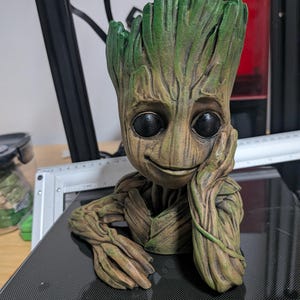 May include: A 3D-printed figure of Groot from Guardians of the Galaxy. The figure has a detailed, textured appearance with a brown and green color scheme. Groot is depicted with a smiling expression, resting his head on his hand.