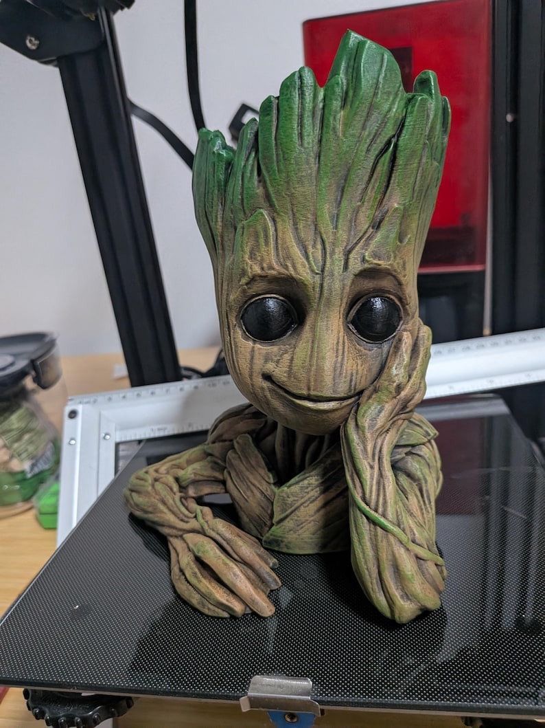 May include: A 3D-printed figure of Groot from Guardians of the Galaxy. The figure has a detailed, textured appearance with a brown and green color scheme. Groot is depicted with a smiling expression, resting his head on his hand.