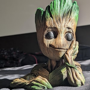 May include: A 3D-printed figure of Groot, a character from Guardians of the Galaxy. The figure has a brown, textured body with green accents on the leaves and head. Groot is depicted with a thoughtful expression, resting his chin on his hand.