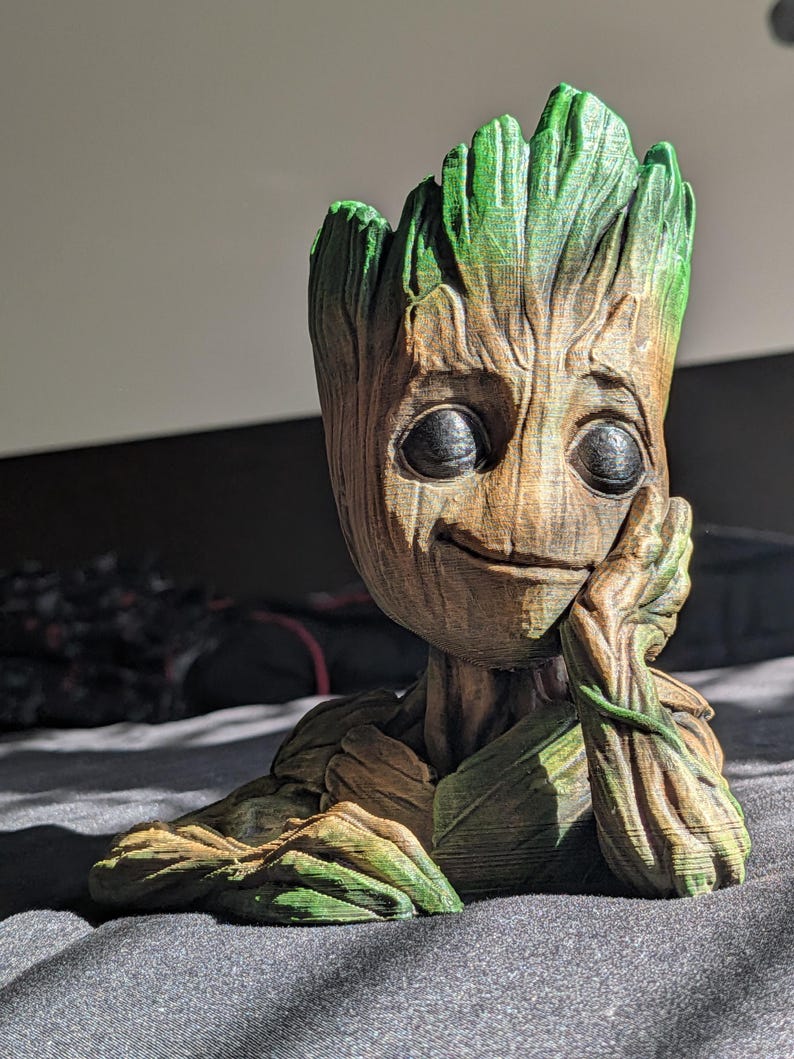 May include: A 3D-printed figure of Groot, a character from Guardians of the Galaxy. The figure has a brown, textured body with green accents on the leaves and head. Groot is depicted with a thoughtful expression, resting his chin on his hand.