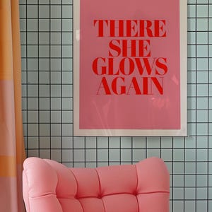 May include: A pink armchair with a tufted back and seat sits in front of a pink and white framed poster with the text "THERE SHE GLOWS AGAIN" in red letters against a light blue tiled wall.