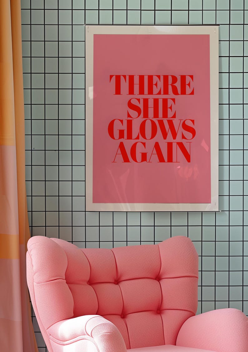 May include: A pink armchair with a tufted back and seat sits in front of a pink and white framed poster with the text "THERE SHE GLOWS AGAIN" in red letters against a light blue tiled wall.