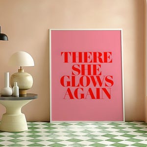 May include: A pink framed poster with the text "THERE SHE GLOWS AGAIN" in red letters.