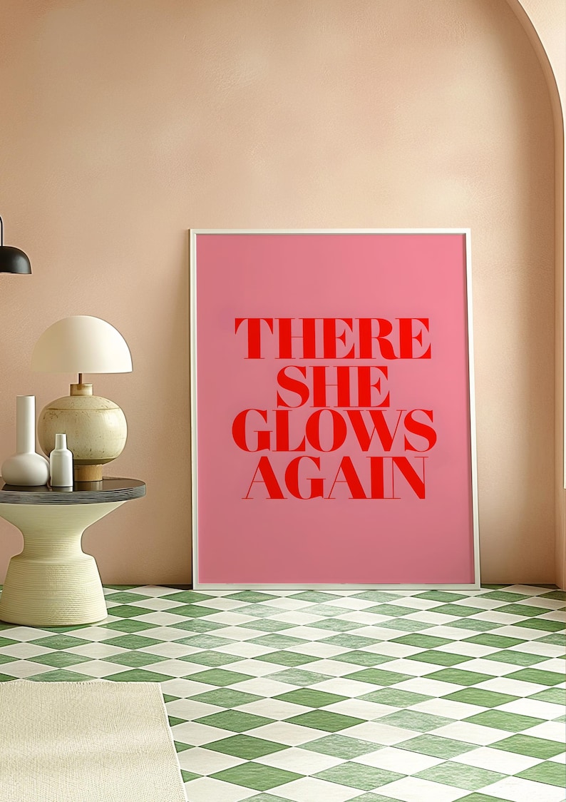 May include: A pink framed poster with the text "THERE SHE GLOWS AGAIN" in red letters.