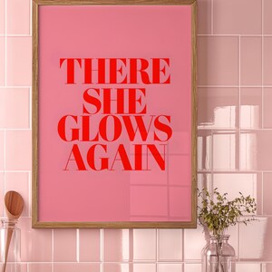 May include: A framed pink poster with the text "THERE SHE GLOWS AGAIN" in red letters.