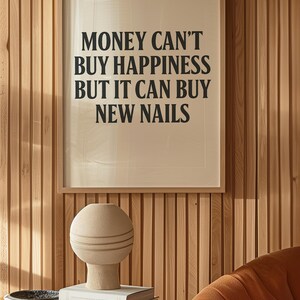May include: A framed print with the text "MONEY CAN'T BUY HAPPINESS BUT IT CAN BUY NEW NAILS" hangs on a wooden wall. A beige vase with horizontal lines sits on a stack of books on a glass table. An orange chair is in the background.
