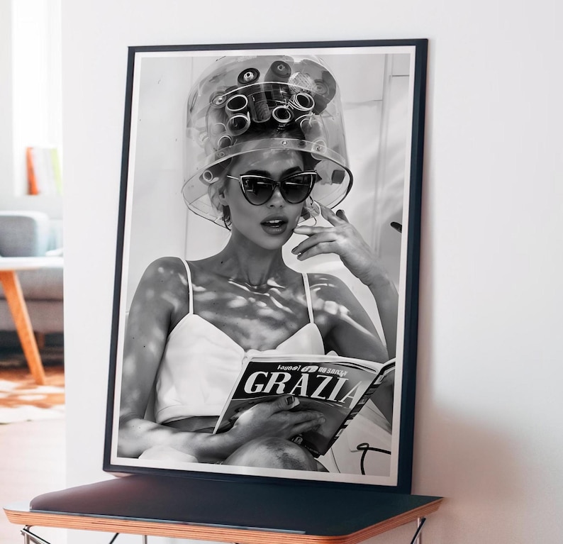 May include: Black and white photo of a woman wearing a hair roller set and sunglasses, reading a magazine called "Grazia".
