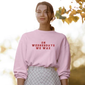 May include: A pink sweatshirt with the text "ON WEDNESDAYS WE WAX" in red lettering. The model is wearing the sweatshirt with a plaid skirt.