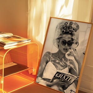 May include: A black and white photo of a woman with hair rollers in her hair, wearing sunglasses and a white tank top, reading a magazine called "GRAZIA".