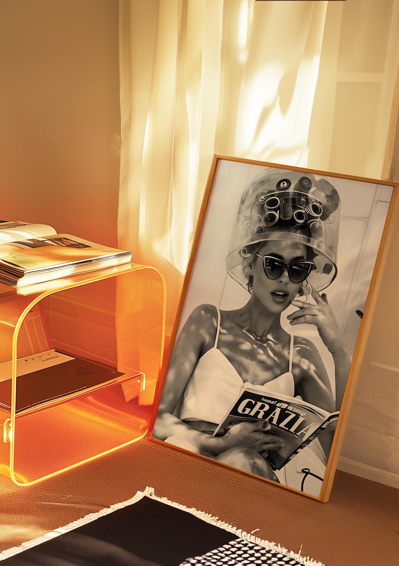May include: A black and white photo of a woman with hair rollers in her hair, wearing sunglasses and a white tank top, reading a magazine called "GRAZIA".