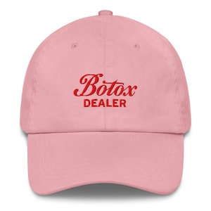 May include: A light pink baseball cap with a curved brim. The cap features the red text "Botox DEALER" embroidered on the front. The cap is made of a soft fabric and has a classic six-panel design.