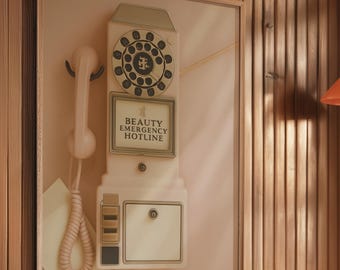 May include: A framed wall art piece featuring a vintage-style telephone in cream and beige tones. The phone has a rotary dial and the text "BEAUTY EMERGENCY HOTLINE". The artwork is set against a light pink background.