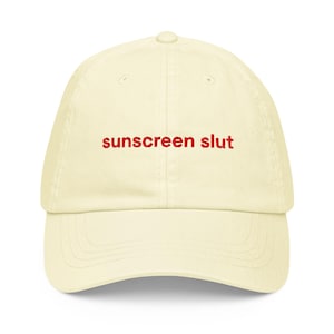 May include: A cream-colored baseball cap with the embroidered text "sunscreen slut" in red. The cap has a curved brim and a six-panel design, suitable for sun protection.