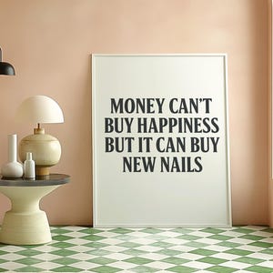 May include: A framed print with the text "MONEY CAN'T BUY HAPPINESS BUT IT CAN BUY NEW NAILS" in black lettering. The print is in a room with a pink wall, a side table with a lamp and vases, and a green and white checkered floor.