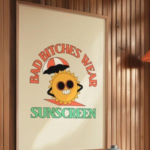 May include: Framed poster with a retro design. The artwork features a smiling sun wearing sunglasses and an umbrella, with the text "BAD BITCHES WEAR SUNSCREEN" in a playful font. The poster is displayed on a wooden wall.