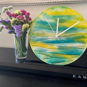 Puede incluir: Un reloj redondo con una esfera blanca y agujas negras. La esfera del reloj está hecha de resina y tiene un diseño abstracto amarillo, verde y azul. El reloj está sobre una superficie negra.