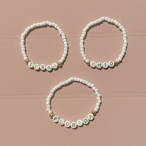 Peut inclure: Trois bracelets de perles avec des perles blanches et des perles de lettres dorées épelant "TEXAS", "OHIO" et "FLORIDA". Les bracelets sont posés sur une surface rose.