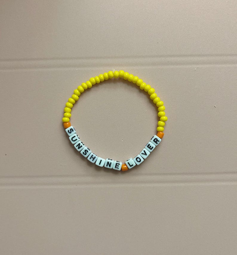 May include: A beaded bracelet with the words "SUNSHINE LOVER" spelled out in white letter beads. The bracelet features yellow and orange beads. The bracelet is a cheerful accessory.