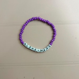 May include: A purple beaded bracelet with the word "ALZHEIMERS" spelled out in white letter beads. The bracelet is made of small, round purple beads. The background is a solid, light pink color.