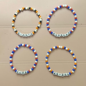Op de afbeelding: Vier armbanden met teamnamen. Eén armband heeft zwarte, oranje en witte kralen met "GIANTS". Een andere heeft rode, witte en blauwe kralen met "DODGERS". De derde heeft rode, witte en blauwe kralen met "CUBS". De laatste heeft oranje, blauwe en witte kralen met "ASTROS".