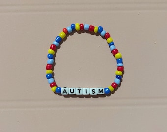 May include: A beaded bracelet with the word "AUTISM" spelled out in white letter beads. The bracelet features a pattern of red, yellow, and blue beads. The bracelet is on a light brown surface.