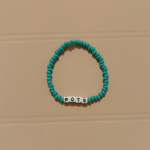 May include: A teal beaded bracelet with the letters P, O, T, and S spelled out in white letter beads. The bracelet is circular and sits on a light brown surface.