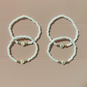May include: Four white beaded bracelets with gold letter charms. The letters spell out "LTBG".
