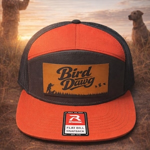 May include: An orange and black flat bill snapback hat with the text "Bird Dawg" on a leather patch. The hat has a mesh back and a Richardson tag. The background shows a hunter, a dog, and flying birds.
