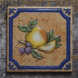 May include: Square ceramic tile with a fruit design. The tile features a yellow and orange apple, a slice of apple, and purple grapes with green leaves. The tile has a blue border with decorative corner accents.